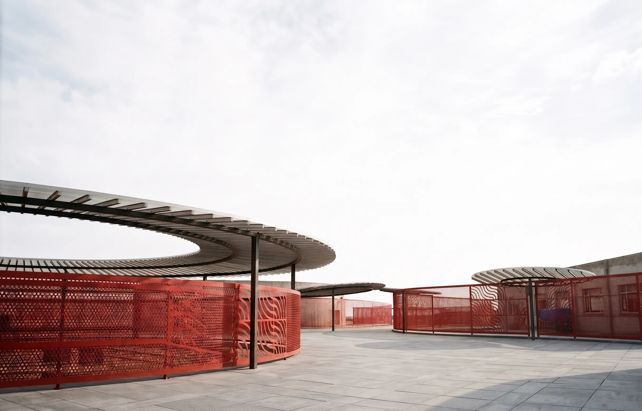 Terraço do colégio com pérgolas circulares e fechamentos vermelhos em chapa perfurada.